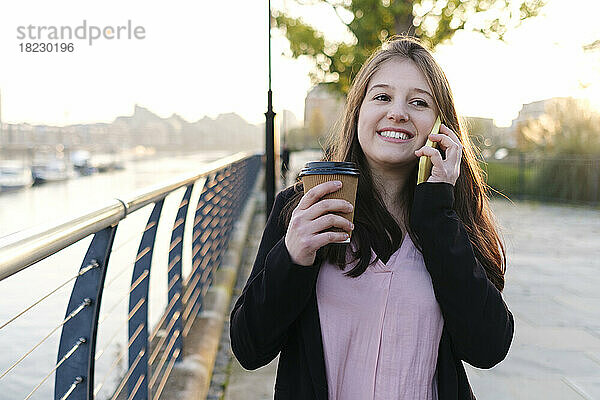 Lächelnde junge Frau hält Einwegbecher in der Hand und telefoniert am Geländer