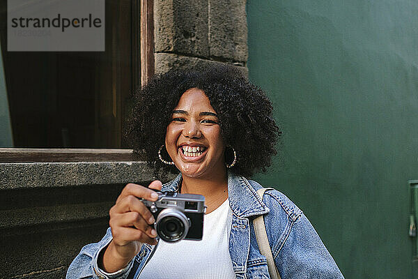 Cheerful young woman with curly hair holding analog camera in front of wall