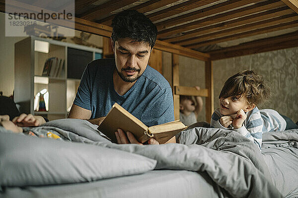Father reads book to his sons on bed at home