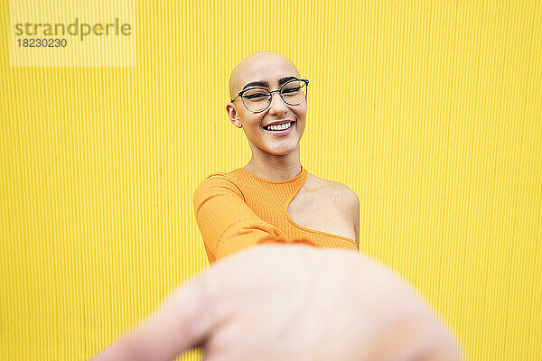 Glückliche Frau mit rasiertem Kopf und Brille vor gelber Wand