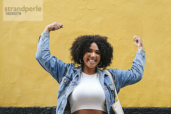 Cheerful young woman flexing muscles in front of wall