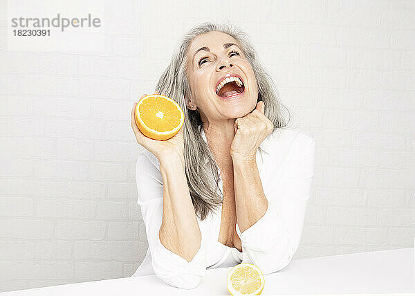 Fröhliche reife Frau mit Orangen sitzt vor der Wand