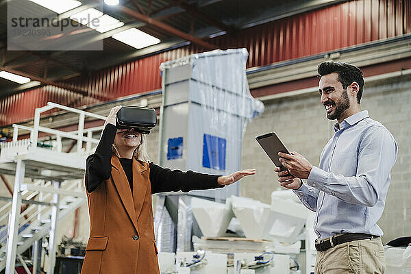 Glücklicher Geschäftsmann mit Kollege  der in der Fabrik einen Virtual-Reality-Simulator trägt