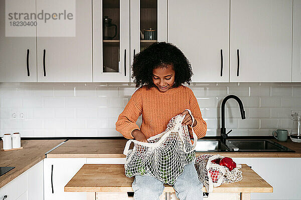 Lächelndes Mädchen  das zu Hause mit einem Netzbeutel voller Gemüse sitzt
