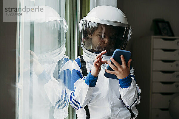 Mädchen im Weltraumkostüm benutzt Smartphone und lehnt zu Hause am Fenster