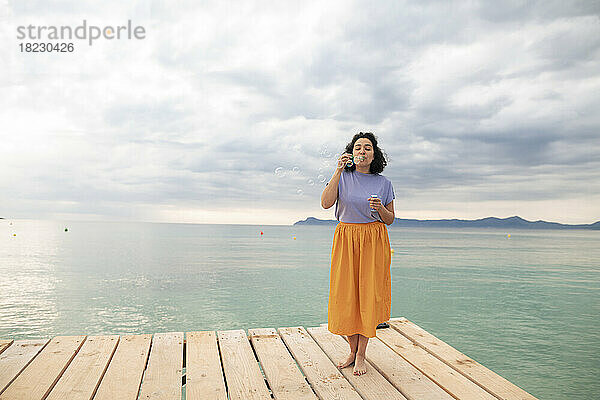 Frau steht auf einem Steg und bläst Seifenblasen vor dem Meer
