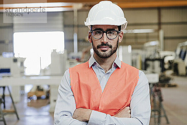 Selbstbewusster Ingenieur mit Brille und Arbeitsschutzkleidung steht mit verschränkten Armen in der Industrie