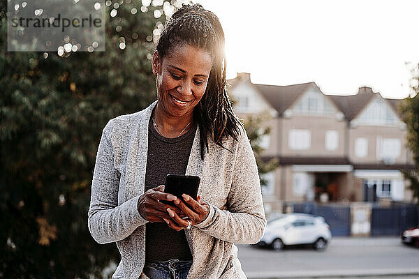 Lächelnde Frau  die bei Sonnenuntergang auf der Terrasse steht und ihr Smartphone benutzt