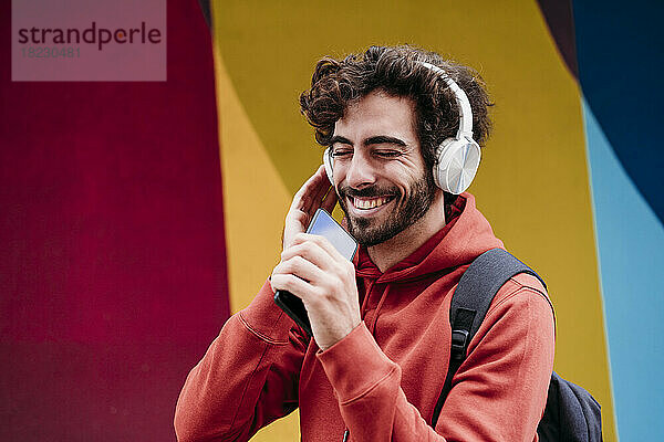 Glücklicher Mann mit geschlossenen Augen  der Musik hört und sein Smartphone vor einer bunten Wand hält