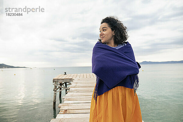Contemplative woman wrapped in shawl standing on jetty