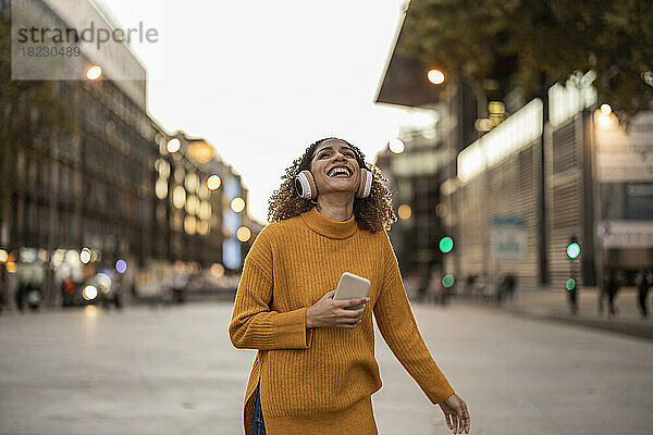 Glückliche junge Frau hält ihr Mobiltelefon in der Hand und genießt Musik  die sie über drahtlose Kopfhörer auf dem Fußweg hört