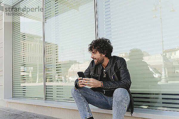 Lächelnder Mann mit Mobiltelefon sitzt auf der Fensterbank