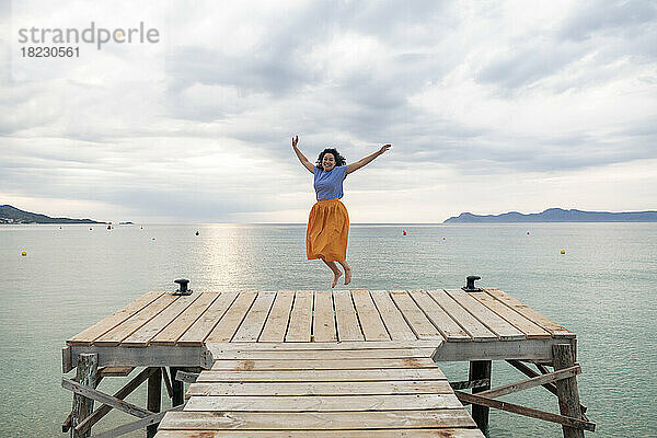 Glückliche Frau springt auf Steg vor dem Meer