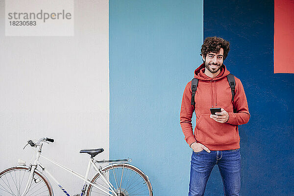 Lächelnder Mann mit der Hand in der Tasche mit dem Fahrrad vor der bunten Wand