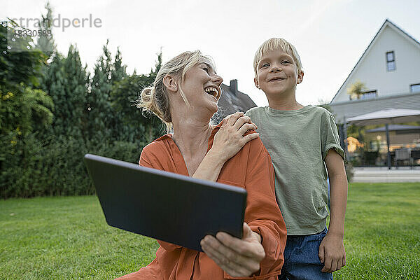 Glückliche Mutter mit Tablet-PC und Blick auf lächelnden Sohn im Garten