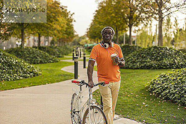 Nachdenklicher älterer Mann hält Einwegbecher und fährt Fahrrad im Park