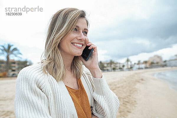 Lächelnde schöne junge blonde Frau  die am Strand mit ihrem Smartphone spricht