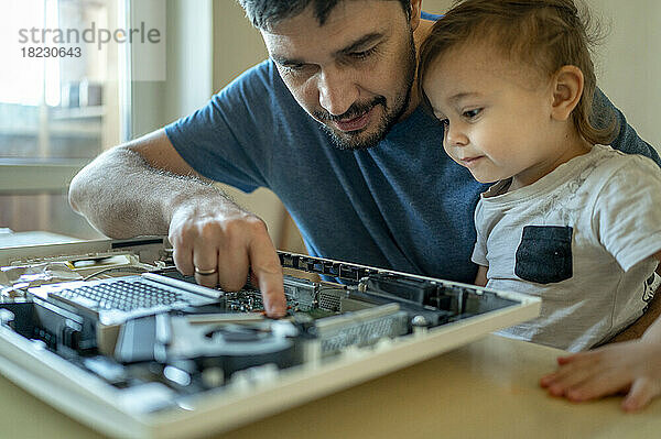 Netter Junge schaut seinem Vater dabei zu  wie er den Computer auf dem Tisch repariert