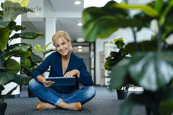 Glückliche Geschäftsfrau mit Tablet-PC sitzt auf dem Boden im Büro
