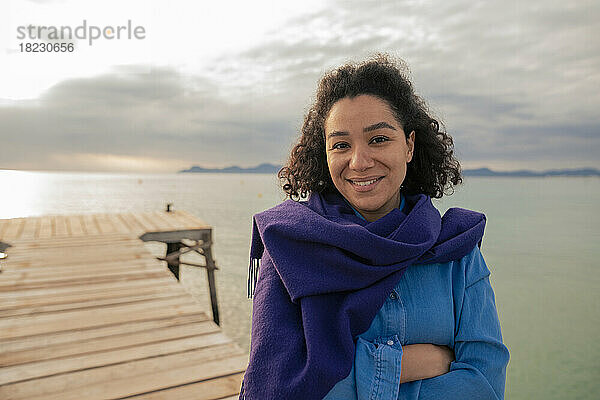 Smiling woman wrapped in shawl standing on jetty at sunset