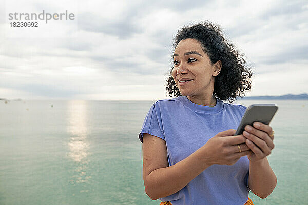 Lächelnde Frau steht mit Smartphone vor dem Meer