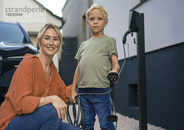 Junge hält Mutter im Vorgarten den Stecker des Ladegeräts für Elektrofahrzeuge in der Hand