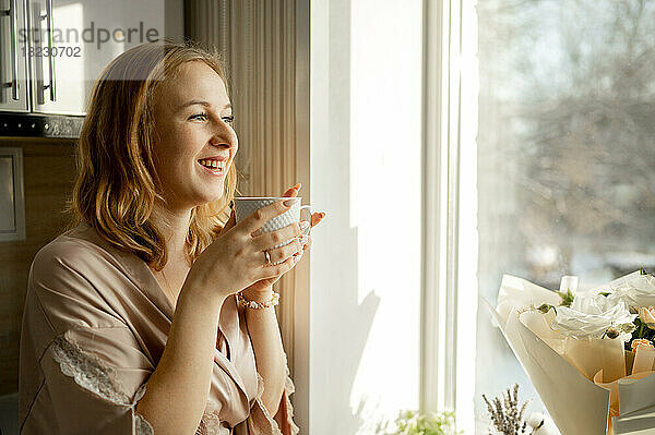Glückliche junge Frau mit Kaffeetasse  die durch das Fenster schaut