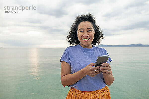 Glückliche Frau steht mit Smartphone vor dem Meer