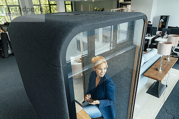 Reife Geschäftsfrau mit Mobiltelefon in schallisolierter Kabine im Büro
