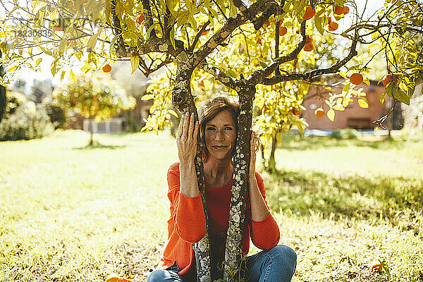 Lächelnde reife Frau sitzt unter einem Orangenfruchtbaum