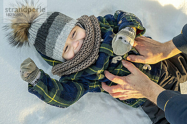 Hände eines Mannes kitzeln einen kleinen Jungen  der auf Schnee liegt