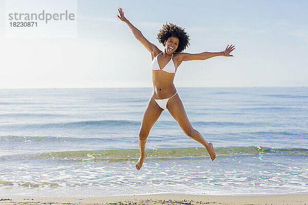 Fröhliche junge Frau springt an einem sonnigen Tag vor das Meer