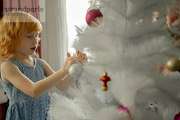 Rothaariges Mädchen schmückt zu Hause künstlichen Weihnachtsbaum