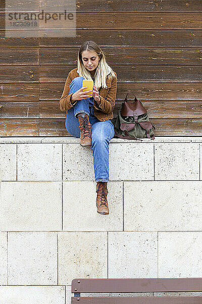 Junge Frau sitzt mit Smartphone an der Wand