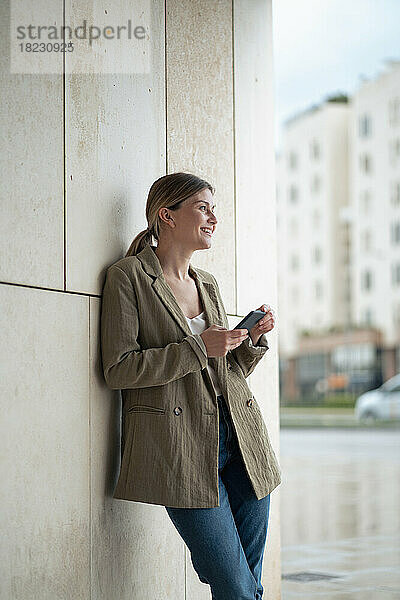 Lächelnde junge Geschäftsfrau mit Mobiltelefon vor der Wand