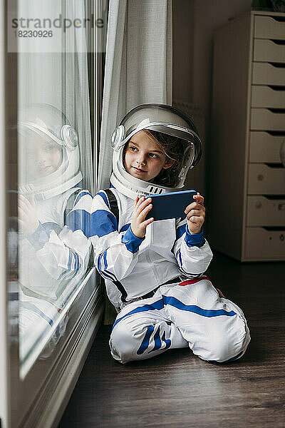 Mädchen im Weltraumkostüm mit Smartphone  das zu Hause durch das Fenster schaut