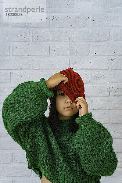 Girl covering eye with knit hat in front of brick wall