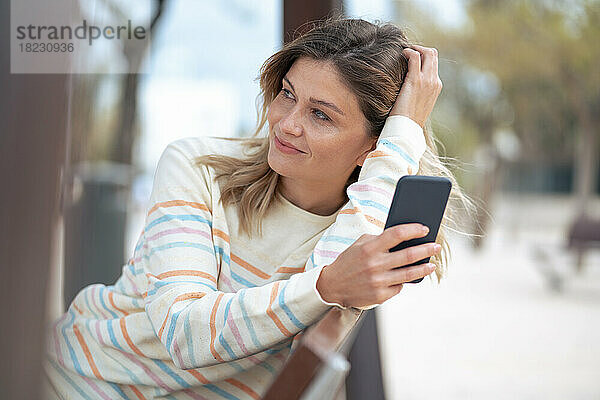 Nachdenkliche junge blonde Frau sitzt mit Smartphone auf einer Bank