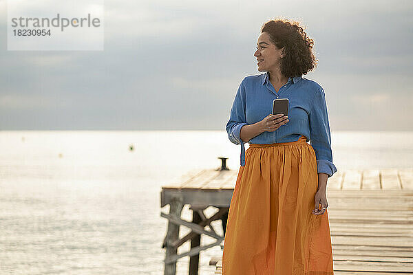Lächelnde Frau steht bei Sonnenuntergang mit Mobiltelefon am Steg