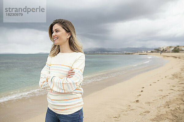 Nachdenklich lächelnde Frau mit verschränkten Armen  die am Strand spaziert