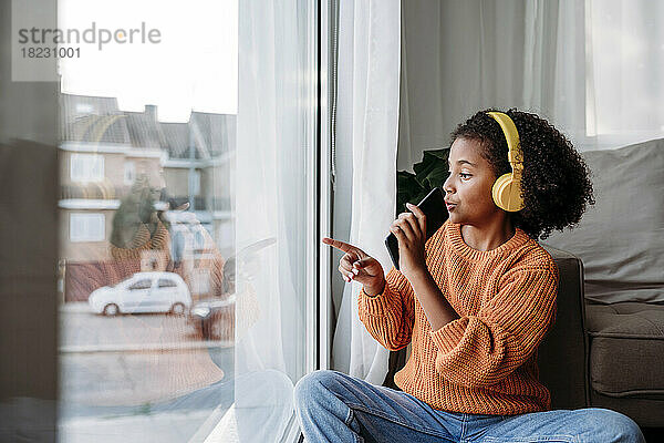 Mädchen mit kabellosen Kopfhörern sitzt mit Smartphone und gestikuliert am Fenster