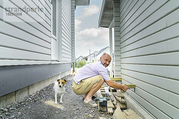 Lächelnder Mann baut Holzwand und kauert neben Hund neben Haus