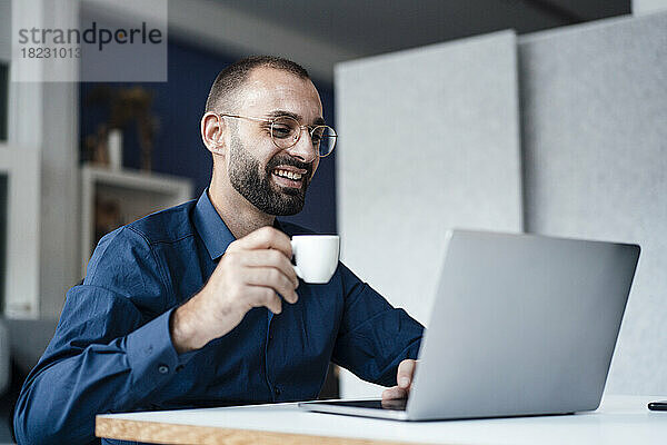 Glücklicher Geschäftsmann hält Kaffeetasse mit Laptop am Schreibtisch im Büro