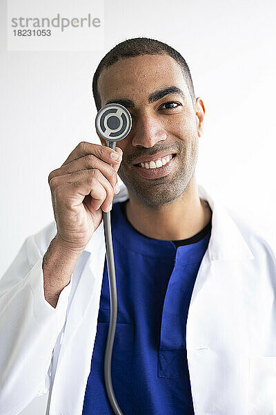 Smiling doctor covering eye with stethoscope against white background