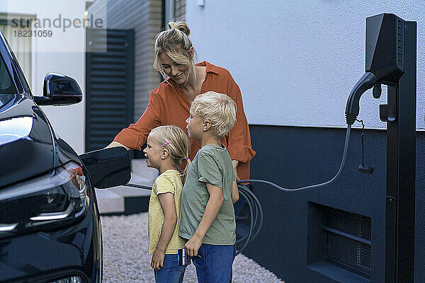 Lächelnde Mutter lädt Kinder im Vorgarten Elektroauto auf