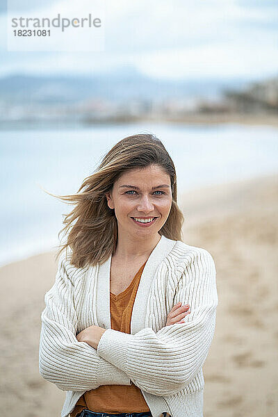 Lächelnde schöne junge Frau mit verschränkten Armen  die am Strand steht