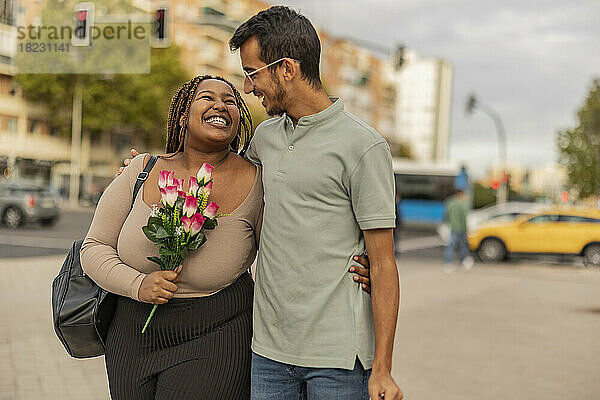 Glückliche junge Frau hält einen Blumenstrauß in der Hand und geht mit Mann am Fußweg spazieren