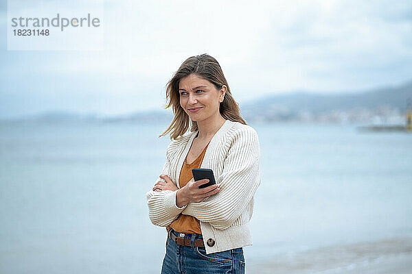 Lächelnde schöne junge Frau mit verschränkten Armen und Smartphone am Strand
