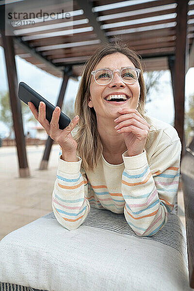 Glückliche junge Frau liegt mit Smartphone auf der Bank
