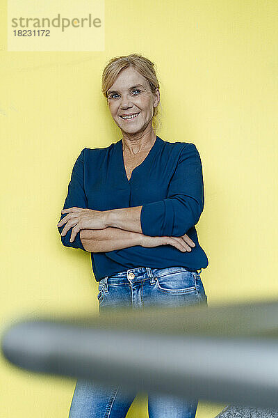 Smiling businesswoman with arms crossed leaning in front of yellow wall